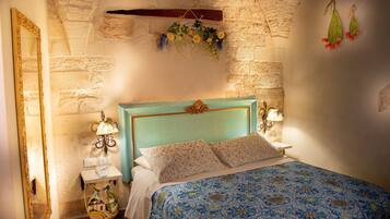 A bedroom with a stone wall, a bed with a blue patterned bedspread, a wooden headboard, and a hanging floral decoration.