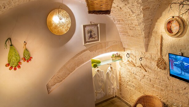 A stone-walled room with a hanging basket, a framed picture, and a television displaying a scene. - Relais Santa Maria (Martina Franca)