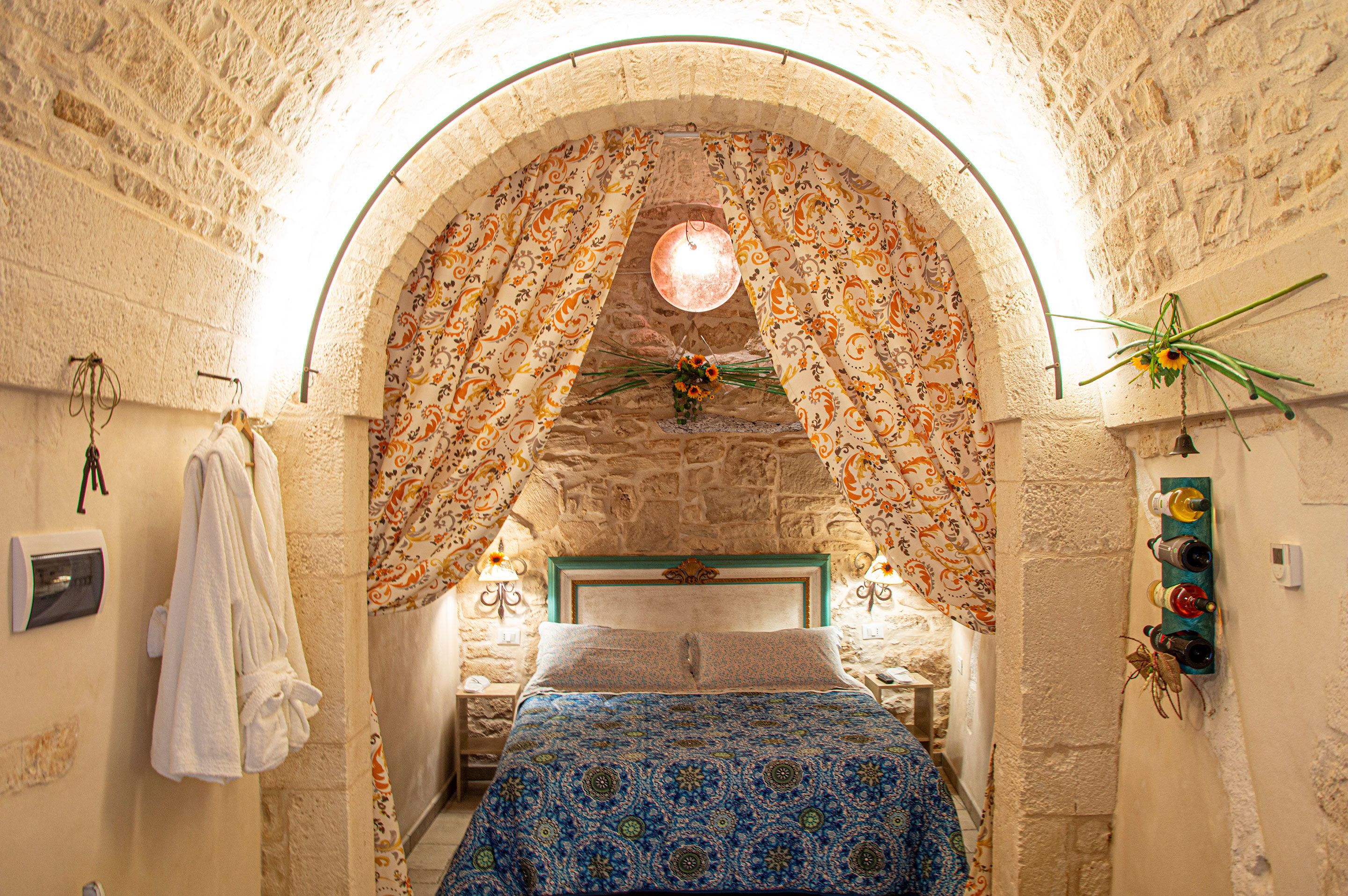 A room with a stone archway, a bed with a patterned bedspread, a hanging flower arrangement, and a wall-mounted wine rack.