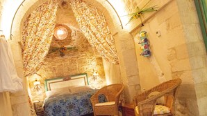 A room with a stone wall, a bed with a patterned bedspread, two wall-mounted lamps, and a wicker chair. - Relais Santa Maria (Martina Franca)