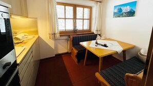 A small, cozy room with a wooden ceiling, a window with curtains, a table with a bottle and papers, a bench, and a radiator.
