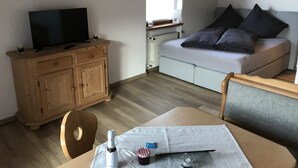 A compact living space with a television on a wooden cabinet, a sofa with cushions, a table with a bottle and glasses, and a window with a view of the outside.
