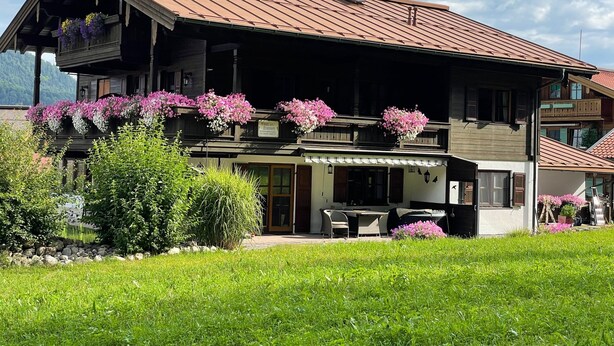 A two-story wooden building with balconies adorned with flowers, a covered outdoor seating area, and a mountain backdrop.