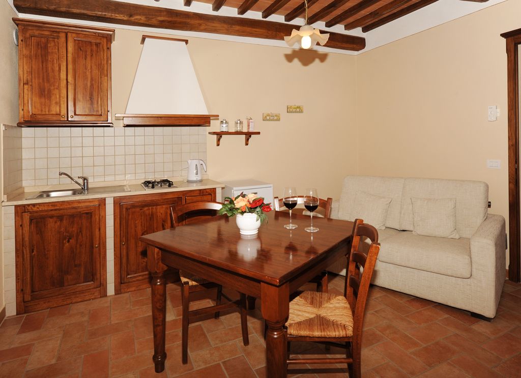 A compact kitchen with wooden cabinets, a tiled backsplash, a dining table with chairs, and a beige sofa in a living area.