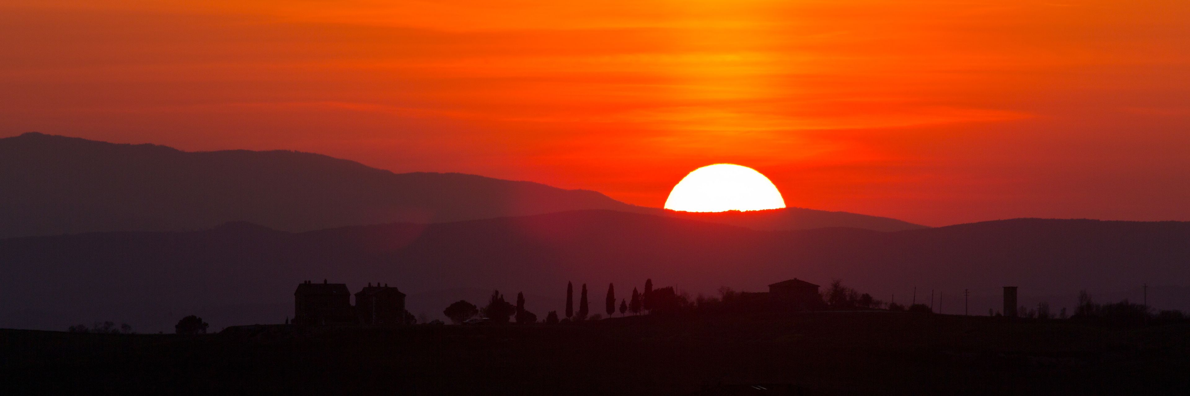 Un atardecer sobre un paisaje montañoso, con siluetas de edificios y árboles.
