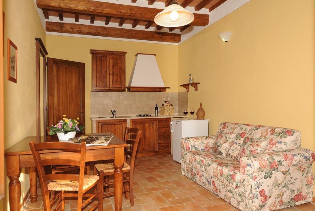 A compact kitchen with wooden cabinets, a dining table with chairs, a floral sofa, and a small refrigerator.
