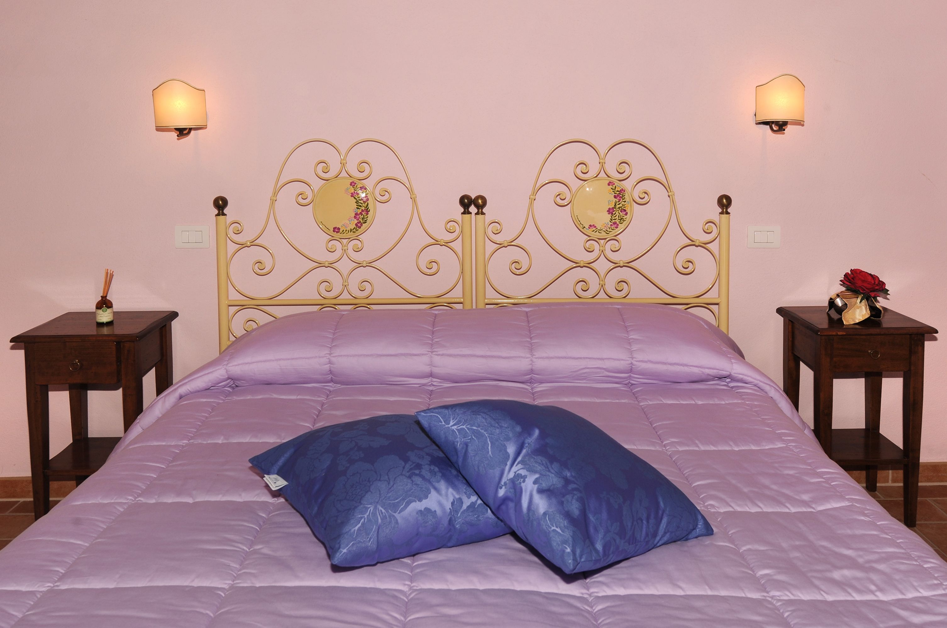 Un lit avec une couette violette et deux oreillers bleus, une tête de lit dorée et décorée, deux tables de chevet avec des lampes et un luminaire fixé au mur.