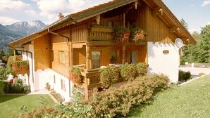 Una cabaña de madera con balcón, rodeada de abundante vegetación y montañas al fondo.