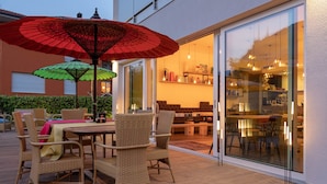 Un patio al aire libre con mobiliario de mimbre, un paraguas rojo y un ventanal de vidrio que da a un comedor interior.