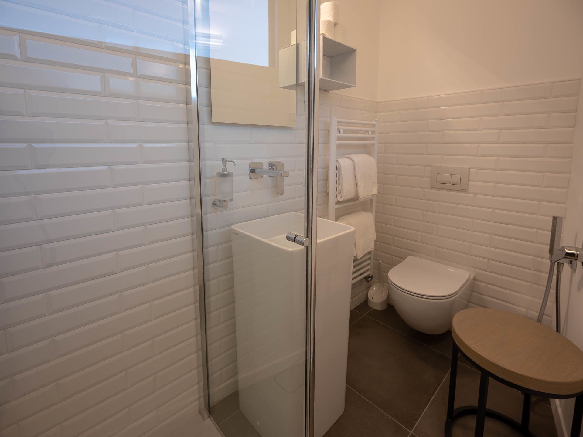 A bathroom with a glass shower enclosure, a toilet, a sink, and a small stool.