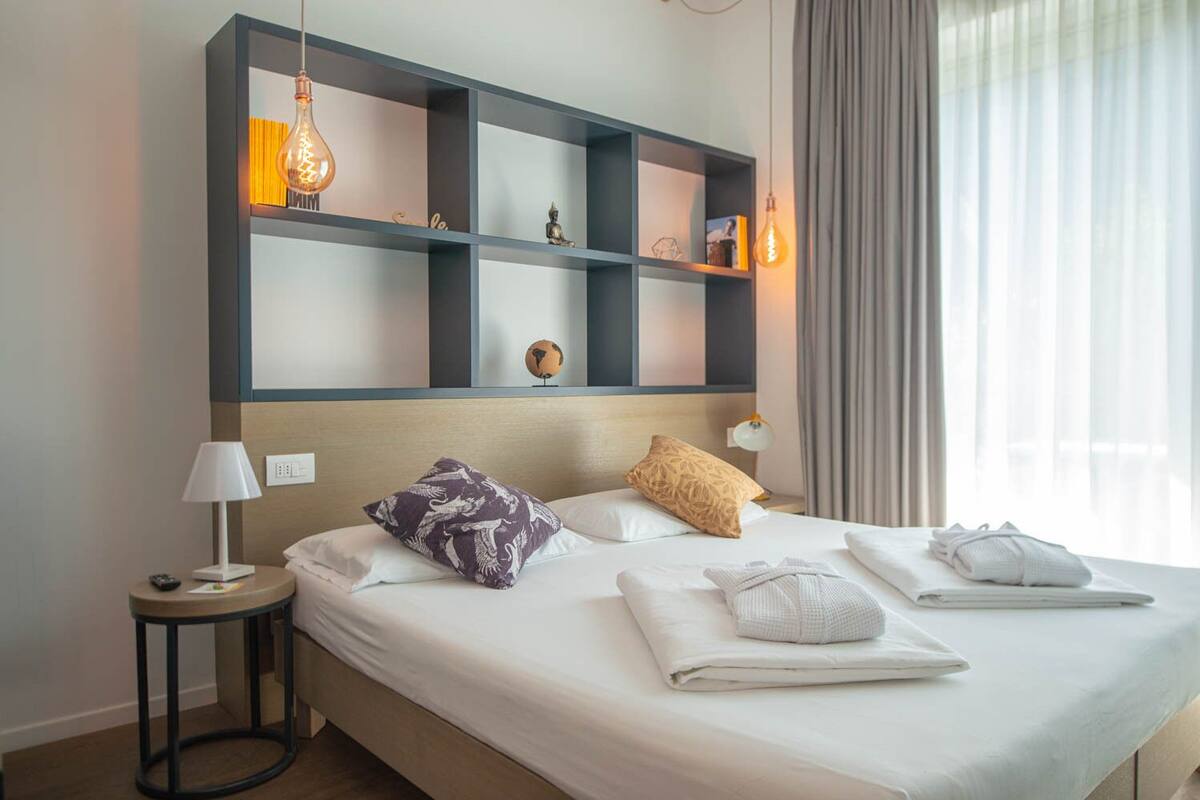 A hotel room with a large bed, two pillows, a bedside table with a lamp, a wall-mounted shelf with decorative items, and a window with curtains.