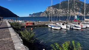 Una marina con barcos amarrados, un sendero de piedra y montañas rocosas al fondo.