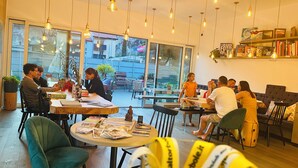 Un café moderno con mesas redondas, lámparas colgantes y grandes ventanales que dan a la calle.