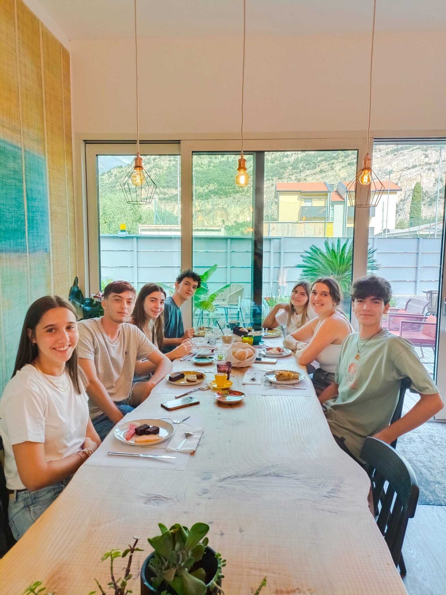 Un grupo de personas sentadas alrededor de una larga mesa de comedor con comida y bebidas, en una habitación con ventanas amplias y lámparas colgantes.