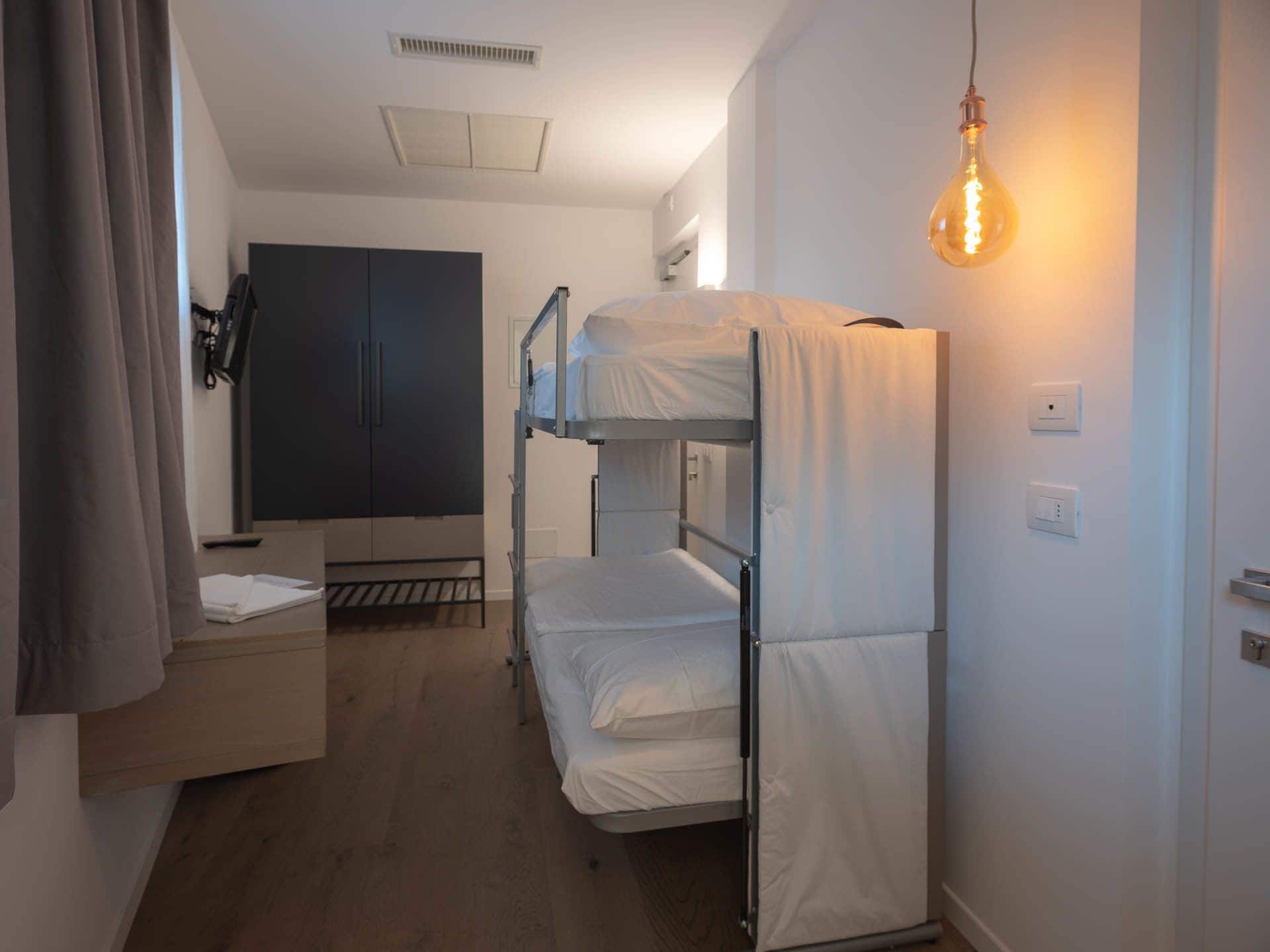 A dormitory room with bunk beds, a wardrobe, a desk, and a hanging light bulb.
