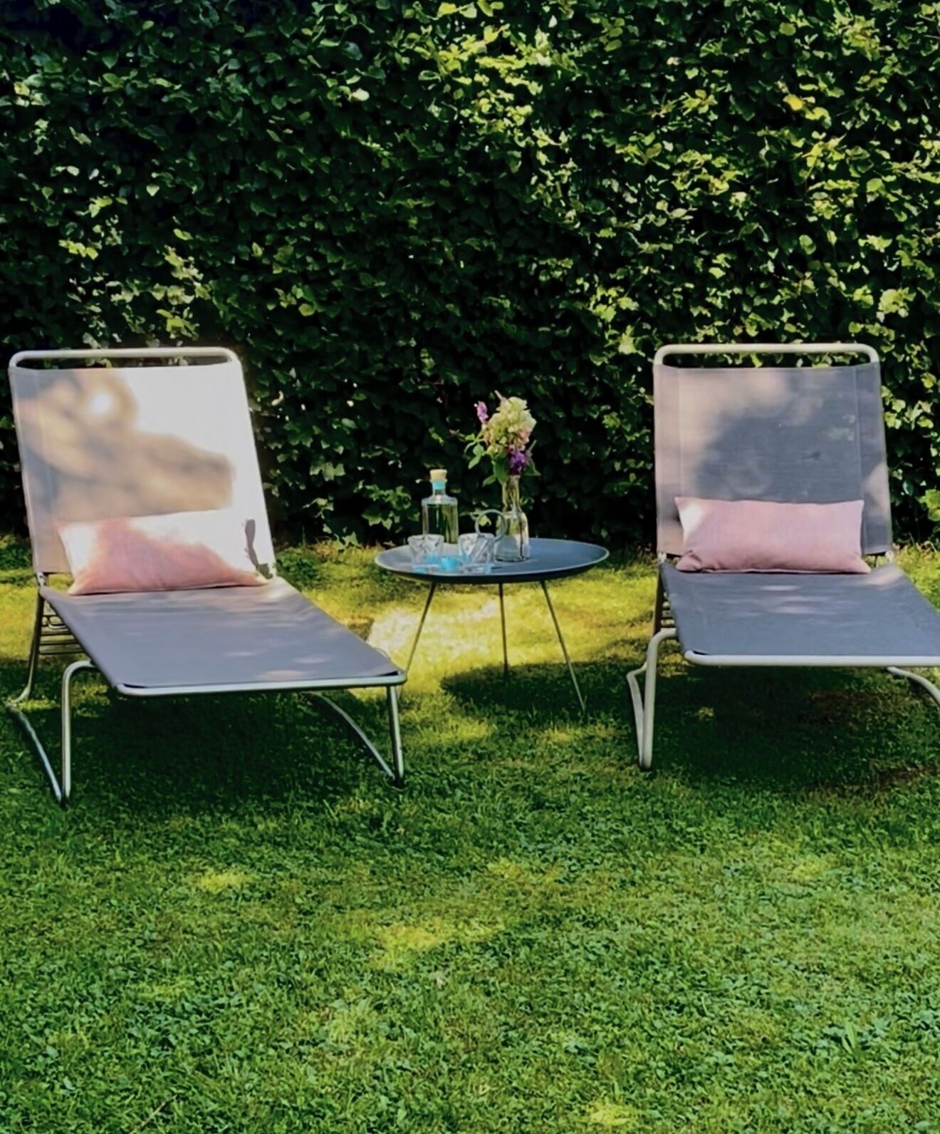 Zwei Lounge-Stühle mit Kissen, ein kleiner runder Tisch mit Blumenvase und ein Hecke im Hintergrund.