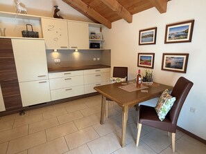A modern kitchen with white cabinets, a wooden dining table, and chairs. There are framed pictures on the wall and a small plant on the counter.