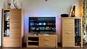 A living room with a flat-screen TV displaying various apps and shows, wooden cabinets with drawers, and a decorative wall light. - Vacation apartment Enzian in Haus Schmid - 90sqm - 1 - 3 persons (Grassau)