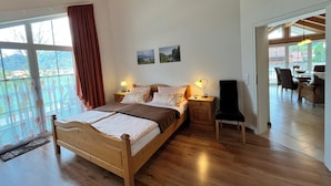 A wooden-framed bed with a nightstand, a lamp, and a chair in a room with wooden floors and a large window. - Vacation apartment Enzian in Haus Schmid - 90sqm - 1 - 3 persons (Grassau)