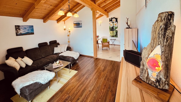 A cozy living room with a wooden ceiling, a sectional sofa, a glass coffee table, and a decorative wooden piece. - Vacation apartment Enzian in Haus Schmid - 90sqm - 1 - 3 persons (Grassau)