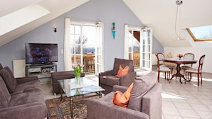 A modern living room with a television, comfortable seating, a glass coffee table, and a view of the outdoors through large windows. - Ferienwohnung Nr. 1 - "Panoramablick", 108 qm Obergeschoss, 1 separates Schlafzimmer (Bad Endorf)
