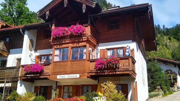 A traditional wooden building with balconies adorned with flowers, a sign reading "Haus Bavaria," and a view of a mountainous landscape. - Zwei-Raum-Ferienwohnung Steinplatte 36qm, Dusche/WC, Extra-Schlafzimmer, Küchenzeile, Balkon (Reit im Winkl)