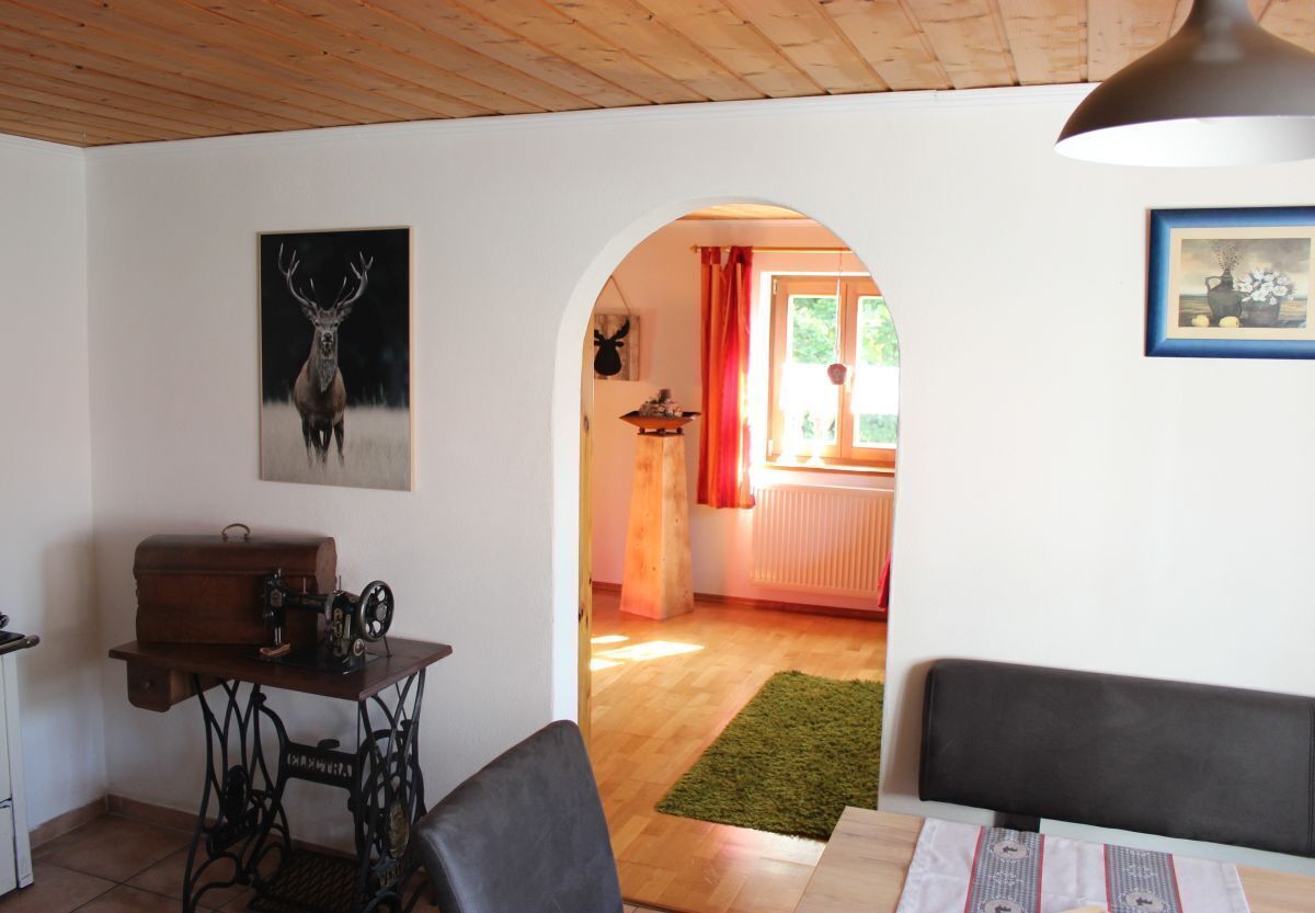Een kamer met een houten plafond, een naaimachine op een tafel, een hertenhof in een lijst, en een deurpost die leidt naar een andere kamer met een radiator en een raam met rode gordijnen.