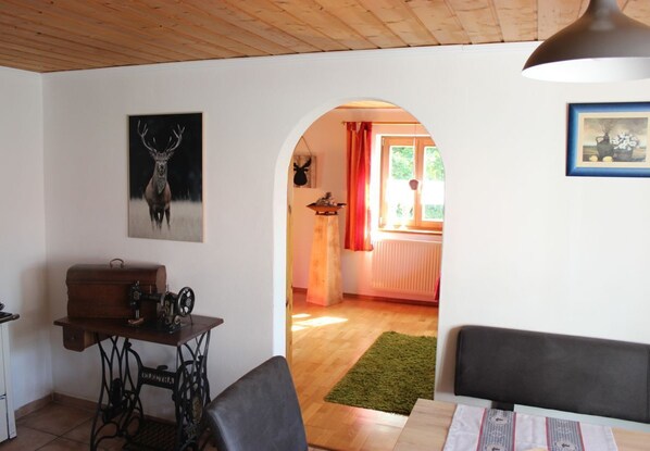 A room with a wooden ceiling, a sewing machine on a table, a framed deer head, and a doorway leading to another room with a radiator and a window with red curtains.