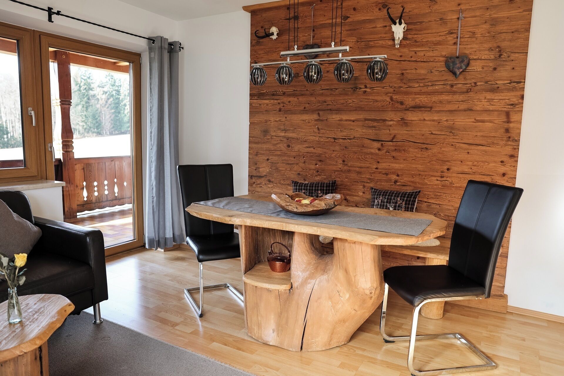 A dining area with a wooden table, black chairs, a wooden wall with mounted lights, and a view through a glass door.
