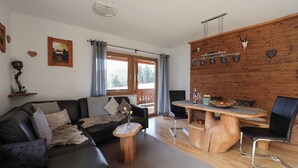 A living room with a sofa, a coffee table, a dining table with chairs, a wooden wall, and a window with curtains. - Vacation apartment Bergfeuer, for 1-4 persons (Berchtesgaden)