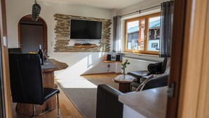 A modern living room with a stone wall, a flat-screen TV, a wooden dining table, and a cozy seating area. - Vacation apartment Bergfeuer, for 1-4 persons (Berchtesgaden)