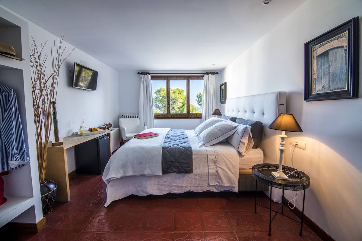 Un dormitorio con cama, mesita de noche, lámpara, televisor, ventana y un escritorio con silla.