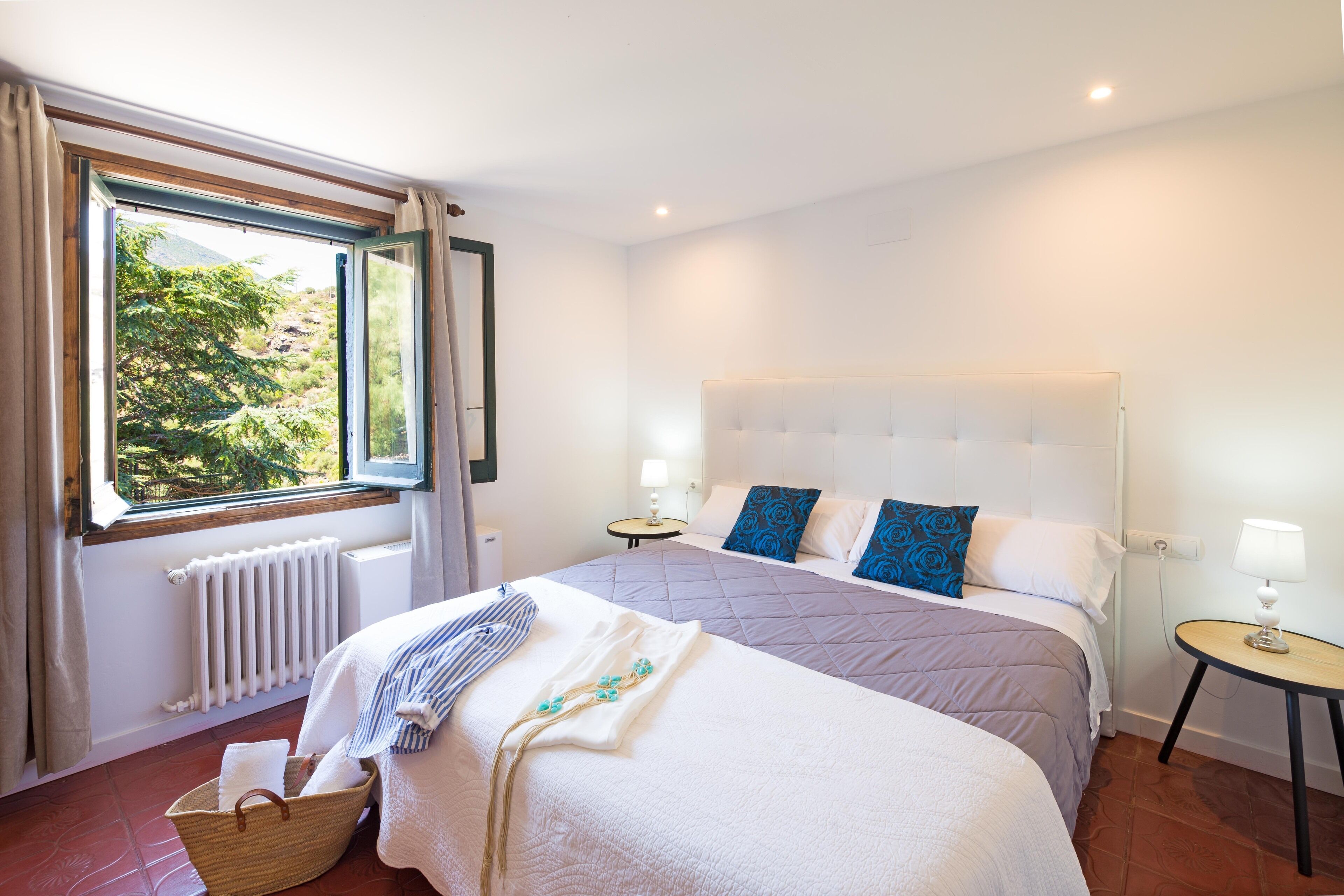 A hotel room with a large bed, a window with curtains, a radiator, a bedside table with a lamp, and a small table with a lamp.