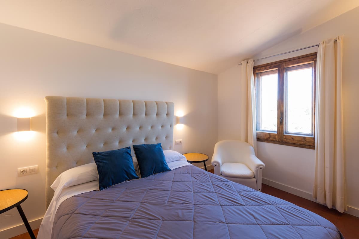 Un dormitorio con una cama grande, una silla, una mesita y un ventanal con cortinas.