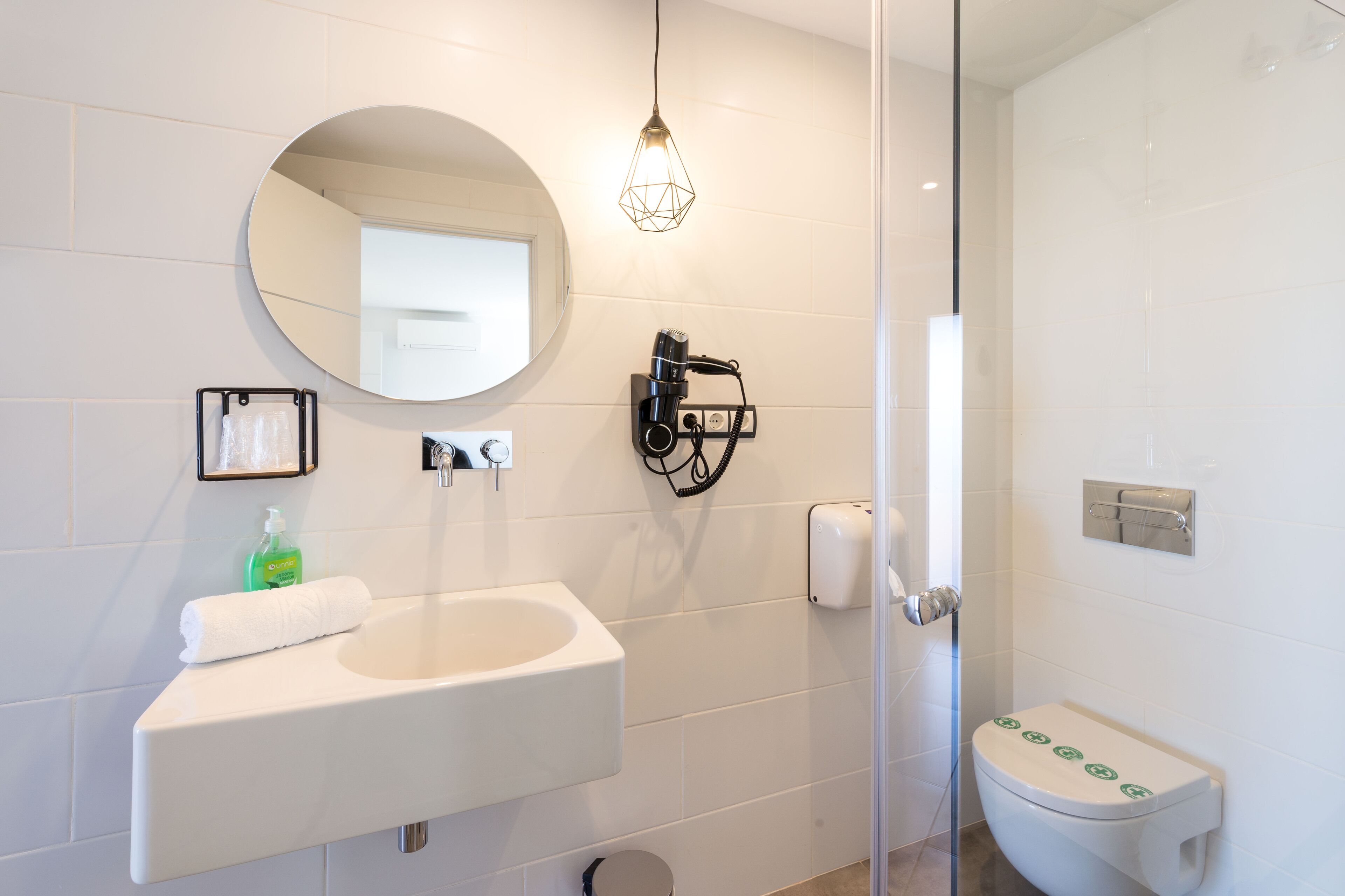 Un baño moderno con un lavamanos blanco, un espejo redondo, un secador de pelo y un inodoro con dispensador de toallas de papel.