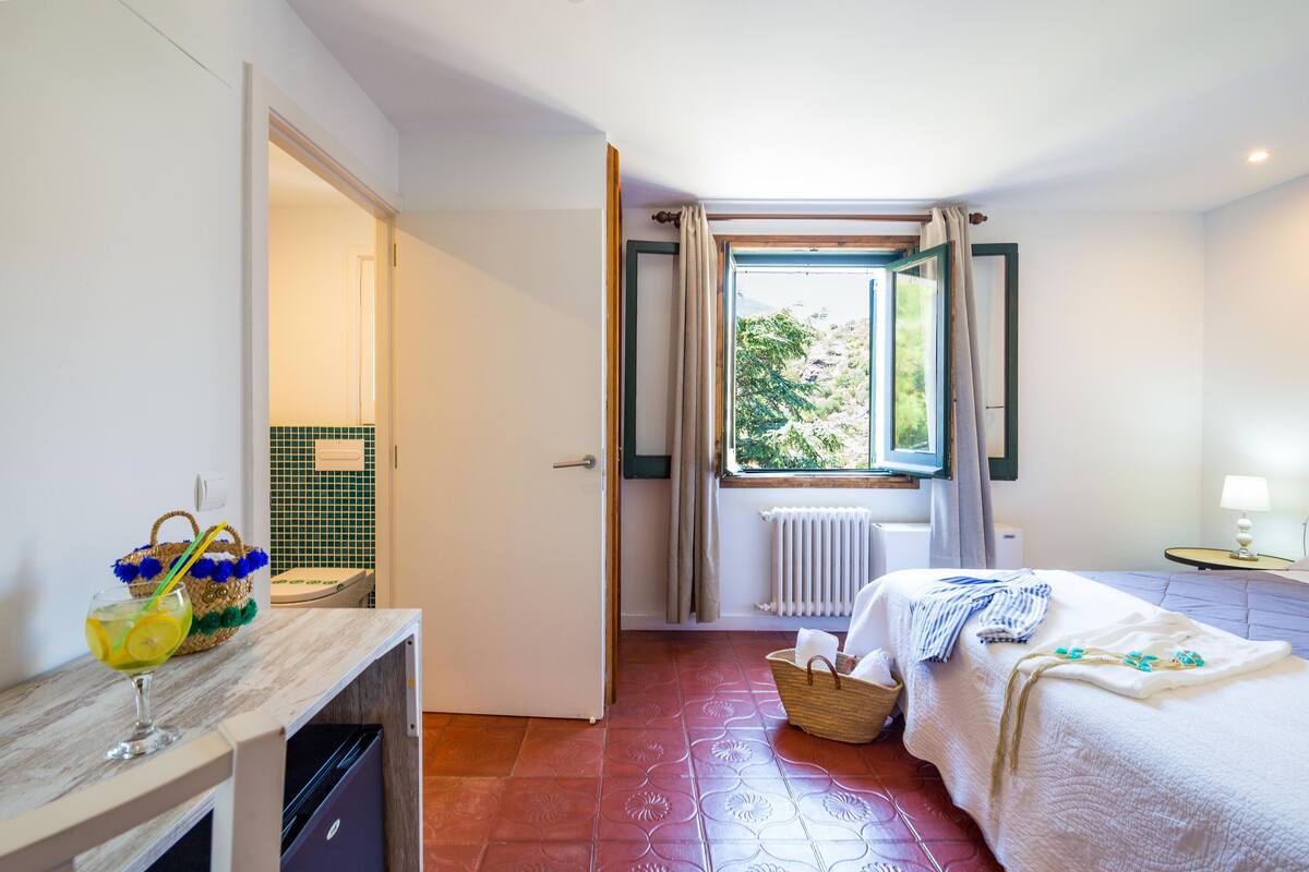 Un dormitorio con cama, ventana con cortinas, radiador y baño con toalla y cesta.