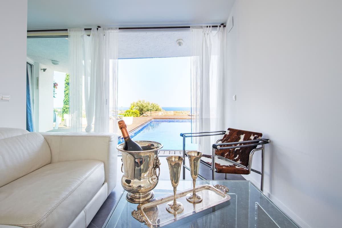 Una sala de estar moderna con un sofá blanco, una mesa de vidrio con cubiertos y vistas a una piscina y vegetación.
