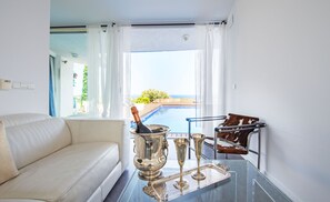 A modern living room with a white sofa, a glass table with silverware, and a view of a pool and greenery outside. - Hotel Rec de Palau (Cadaqués)