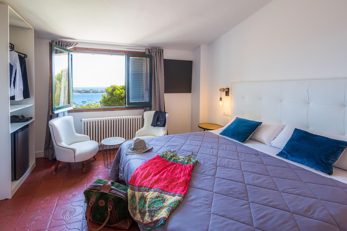 Una habitación de hotel con una cama, una silla, una mesa, una maleta y vistas al mar.