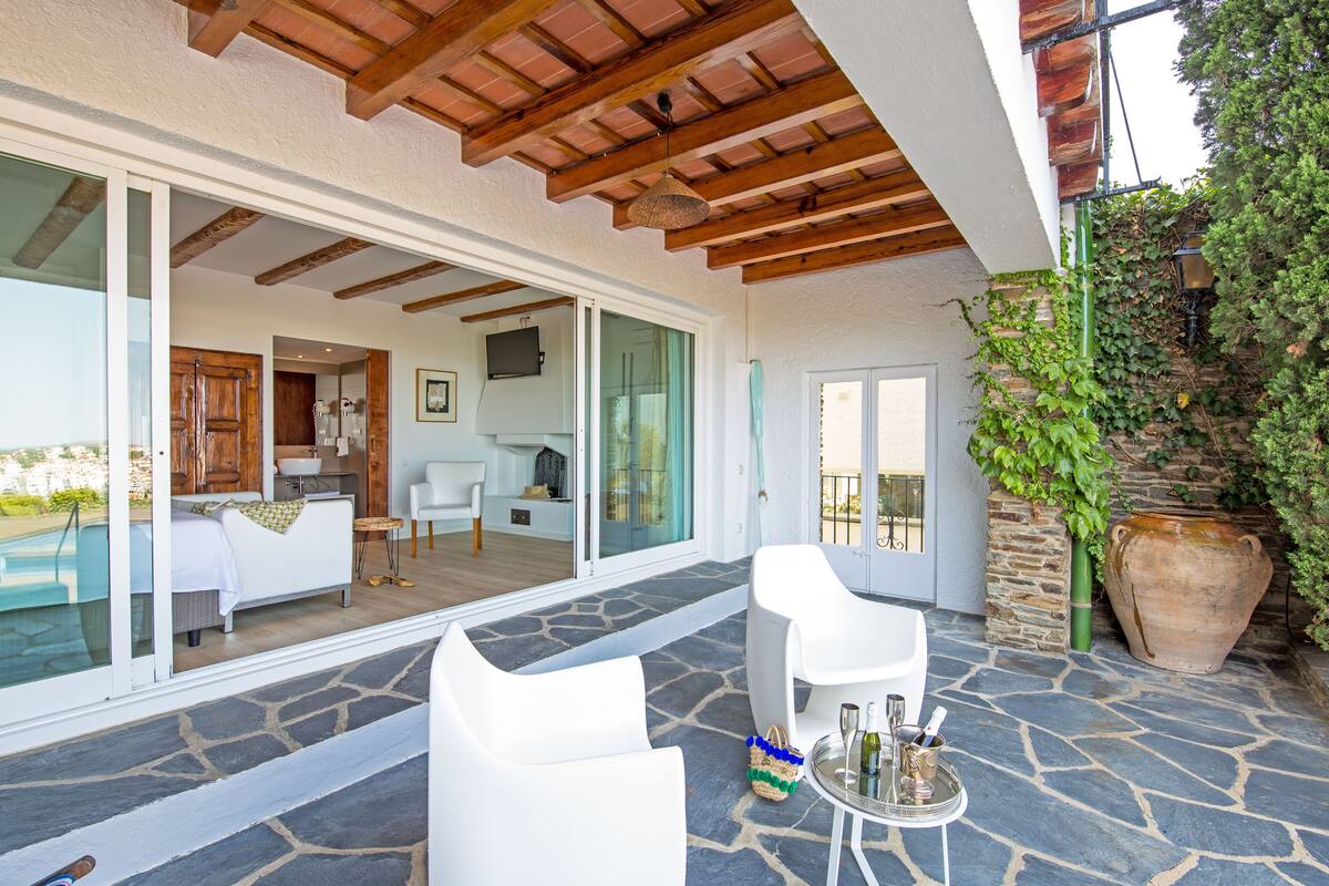 Un espacio exterior moderno con suelo de piedra, mobiliario blanco y vistas a la piscina y la cocina.