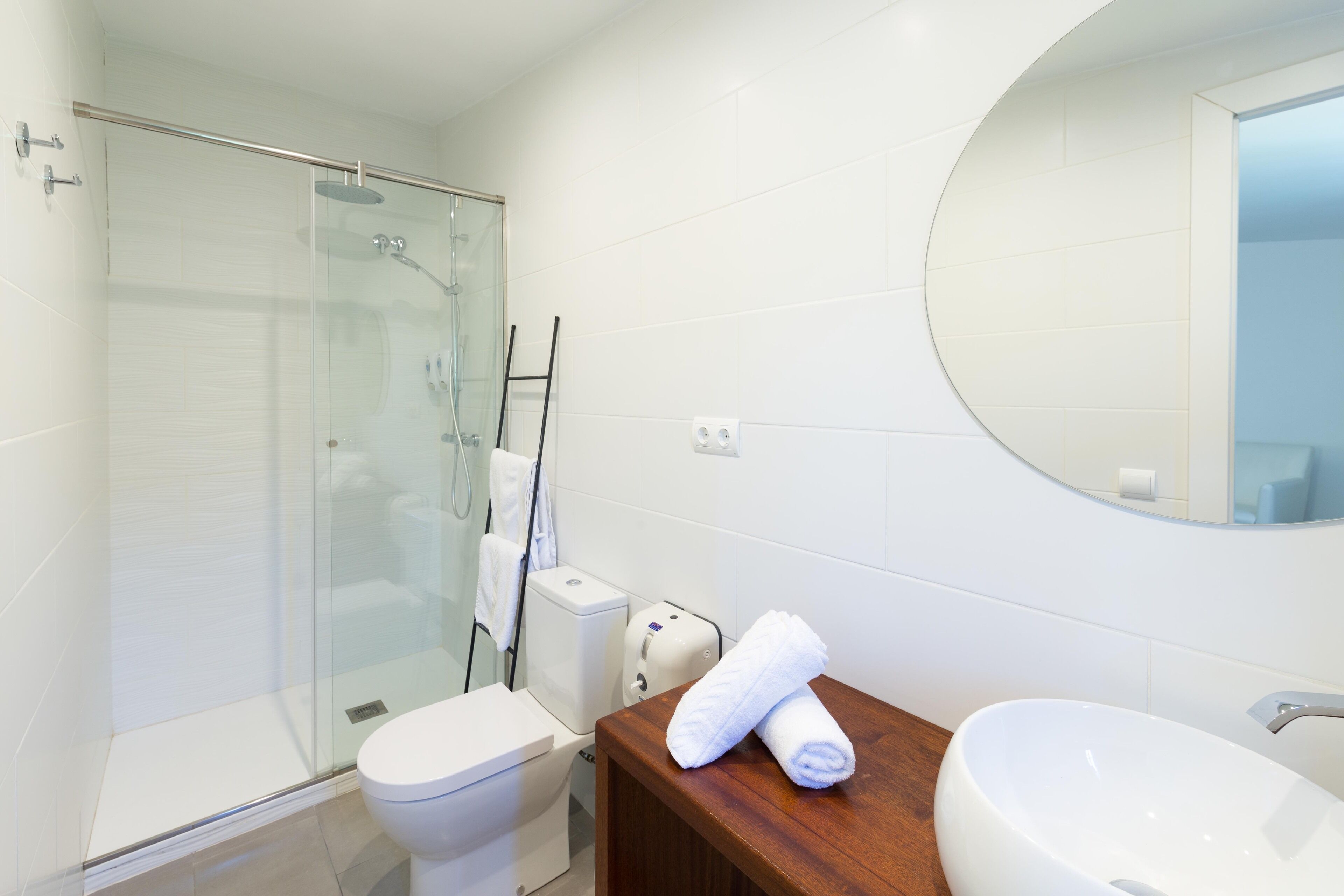 Un baño moderno con ducha de vidrio, inodoro blanco, tocador de madera con lavamanos y espejo.