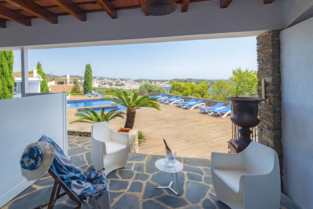 Un patio con sillas blancas, una mesa pequeña con una botella y vasos, y una vista de una piscina y un pueblo al fondo.