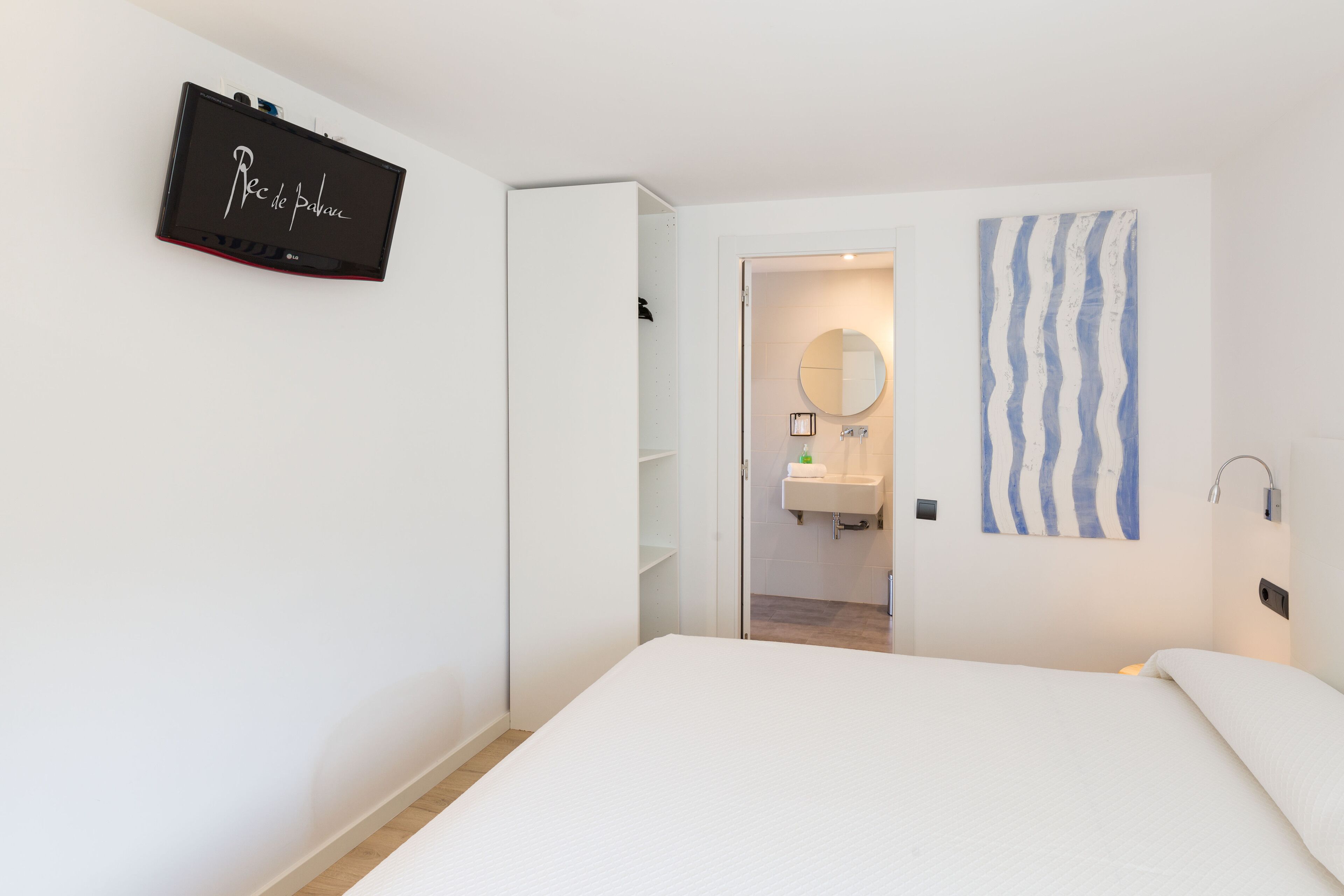 Habitació d'hotel amb llit, televisió penjada a la paret, bany amb rentaplat i mirall, i una obra d'art amb disseny de ratlles.