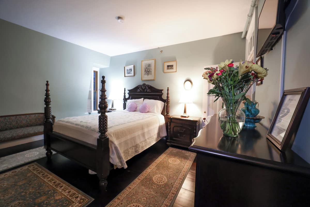 Un dormitorio con una cama con dosel, una mesita de noche con lámpara, un florero con flores y cuadros en la pared.