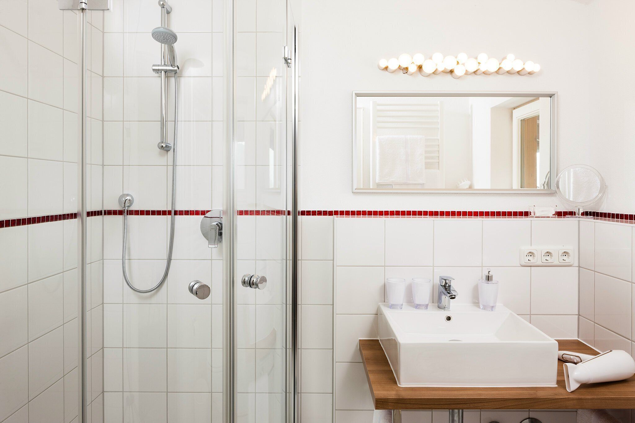 A modern bathroom with a glass-enclosed shower, a white sink with a wooden countertop, and a mirror with lights above it.