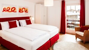 Een tweepersoonsbed met witte en rode beddengoed, een rood hoofdeinde en een groot raam met rode gordijnen.