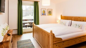 Een houten bed met witte lakens, een groen gordijn, een raam met uitzicht en twee ingelijste foto's aan de muur.