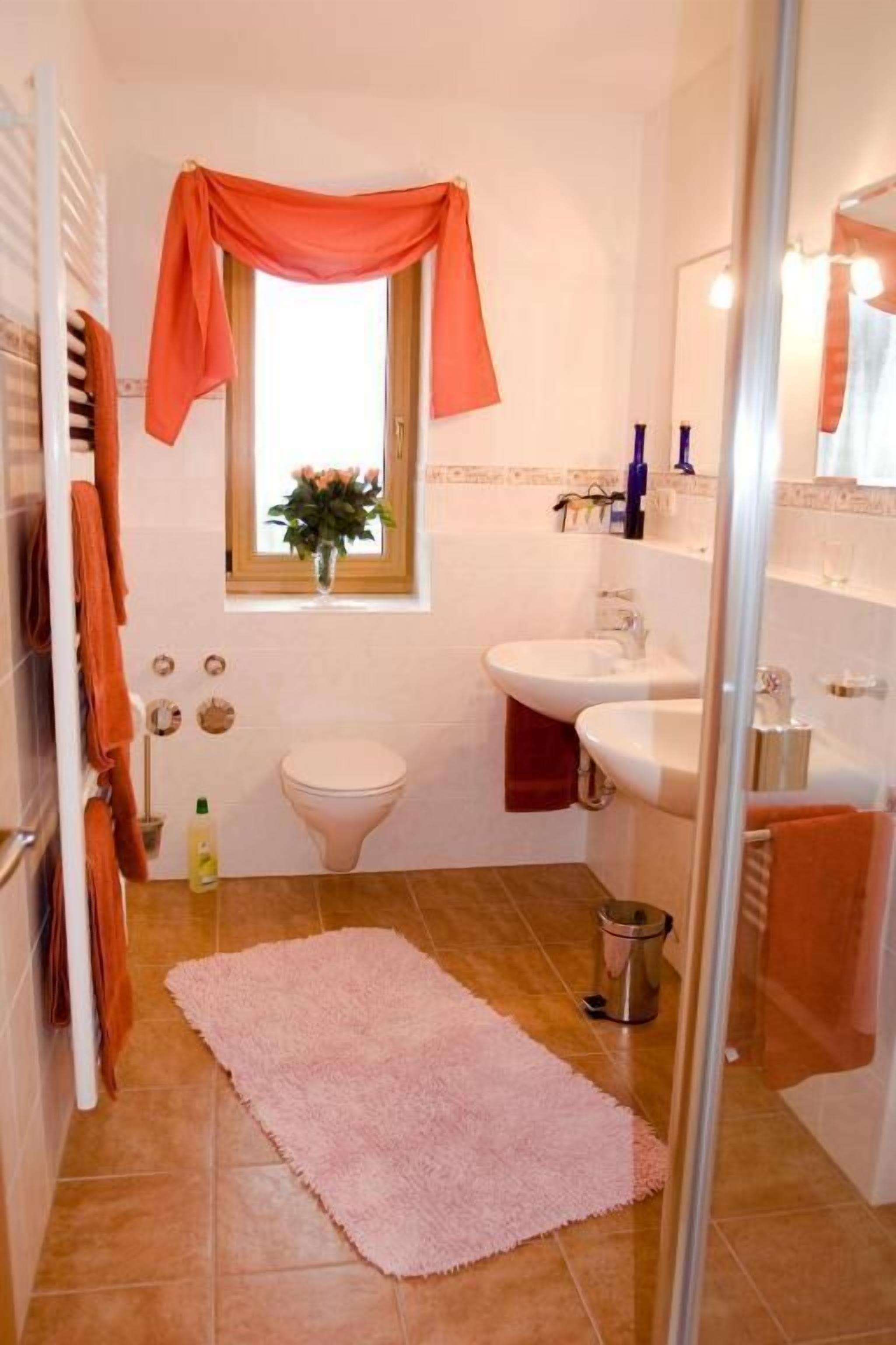Ein Badezimmer mit Toilette, zwei Waschbecken, einem Fenster mit Vorhang, einem rosa Teppich und einem Spiegel.