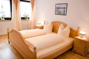 Uma cama de casal com estrutura de madeira, duas mesas de cabeceira com luminárias, uma janela com cortinas e um quadro na parede.