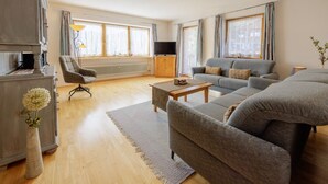 A spacious living room with a grey sofa set, a wooden coffee table, and a large window with curtains.